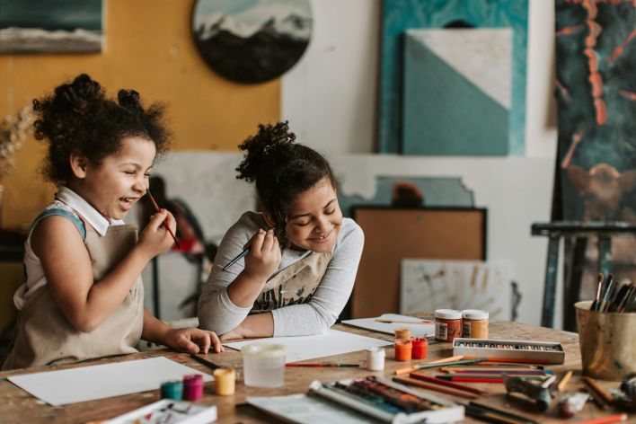 Two young girls happily painting during an indoor art workshop, expressing creativity and learning.