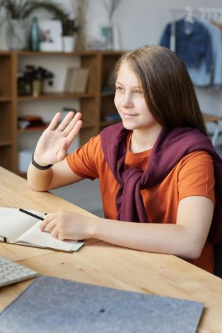 A teenage girl waves at her computer during an online learning session, sitting at a wooden desk indoors.