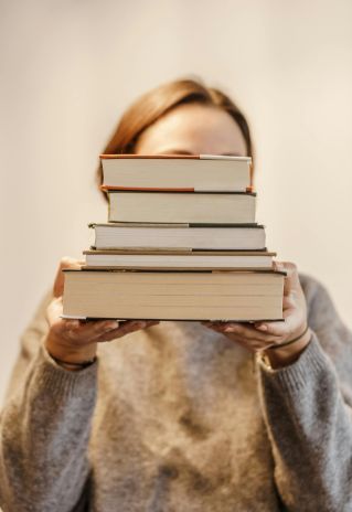 Anonymous woman in soft sweater covering face with pile of books on light background